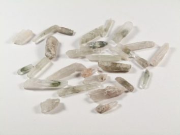 Aktynolit - kamień surowy, nieszlifowany, w kwarcu. Kilka małych kryształków aktynolitu w kwarcu leży rozrzuconych na białym tle.