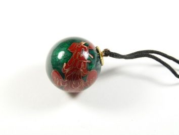 Zielona kulka z czerwonym wzorem, zawieszona na czarnym sznurku, leży na białym tle.
