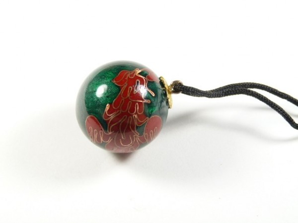 Zielona kulka z czerwonym wzorem, zawieszona na czarnym sznurku, leży na białym tle.