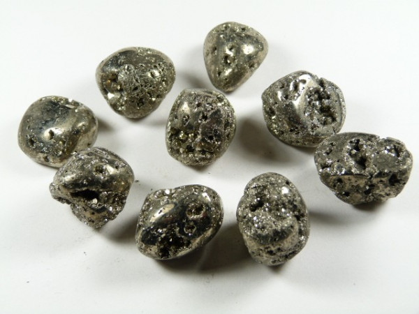Piryt - kamień gładzony, polerowany, szlifowany i bębnowany w formie otoczaka. Dziewięć bryłek pirytu o metalicznym połysku ułożonych na białym tle.
