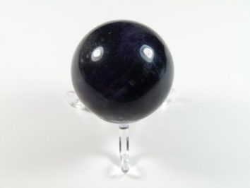 Kula fluoryt szlifowany na podstawce. Ciemnofioletowa kula z minerału na przezroczystej podstawce, na białym tle.