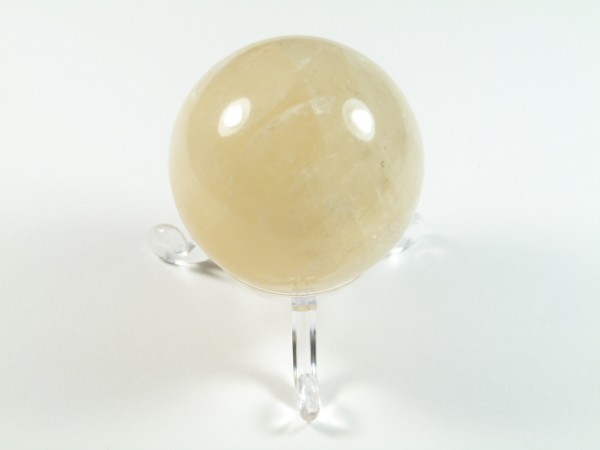 Kula kalcyt złoty szlifowany o średnicy 4,5 cm. Jasna kula z kamienia na przezroczystej podstawce, ustawiona na białym tle.