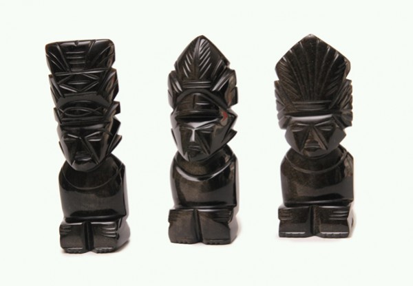 Trzy czarne, rzeźbione figurki siedzących postaci o geometrycznych twarzach i ozdobnych nakryciach głowy.