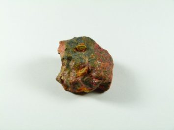 Orpiment/ realgar - kamień naturalny, surowy, nieszlifowany. Nieregularny kamień o czerwono-zielonej barwie leży na białym tle.