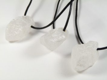 Trzy naszyjniki z surowymi białymi kamieniami na czarnych sznurkach, leżące na białym tle.