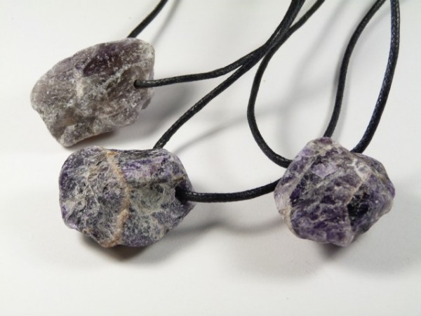 Trzy surowe kamienie ametystu na czarnych sznurkach, ułożone na białym tle.