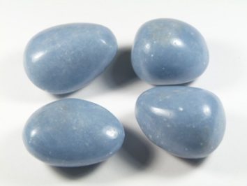 Angelit - kamień gładzony, polerowany, szlifowany i bębnowany w formie otoczaka. Cztery gładkie, niebieskie kamienie leżą na białym tle.