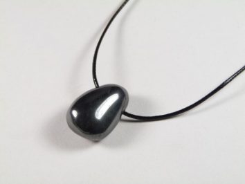 Wisiorek hematyt gładzony z dziurką na sznureczku. Czarny, błyszczący kamień na czarnym sznurku, naszyjnik leży na białym tle.