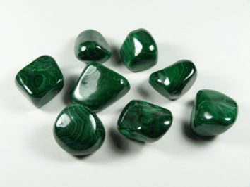 Malachit - kamień gładzony, polerowany, szlifowany i bębnowany w formie otoczaka. Siedem zielonych, oszlifowanych kamieni malachitu na białym tle.