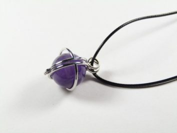 Wisiorek z fioletowym kamieniem oplecionym drutem na czarnym rzemyku.