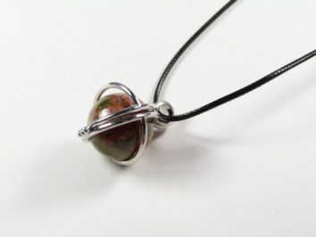 Wisiorek z kamieniem w srebrnej oprawie na czarnym sznurku, leżący na białym tle.