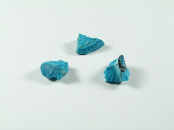 Chryzokola. Trzy małe, nieoszlifowane kawałki turkusu na jasnym tle.
