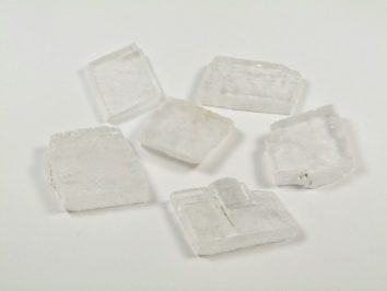 Spar islandzki - kamień naturalny, surowy, nieszlifowany. Sześć przezroczystych kryształów leży na białym tle.