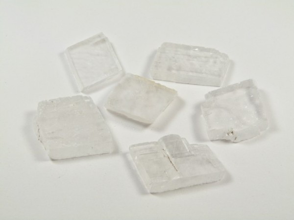 Spar islandzki - kamień naturalny, surowy, nieszlifowany. Sześć przezroczystych kryształów leży na białym tle.