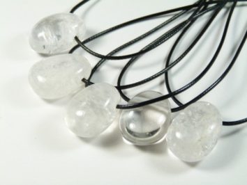 Kryształ górski szlifowany z dziurką, wisiorek na sznureczku. Pięć przejrzystych kamieni na czarnych sznurkach, ułożonych na białym tle.