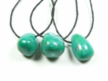 Wisiorek howlit chryzokola gładzony z dziurką na sznureczku. Trzy zielone kamienie na czarnych sznurkach, ułożone obok siebie na białym tle.