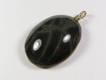 Owalny wisiorek z ciemnego kamienia z jasnymi żyłkami, oprawiony w metalową ramkę.