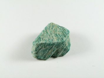 Amazonit - kamień naturalny, surowy, nieszlifowany. Zielony minerał amazonit na białym tle, widoczna nieregularna struktura skały.