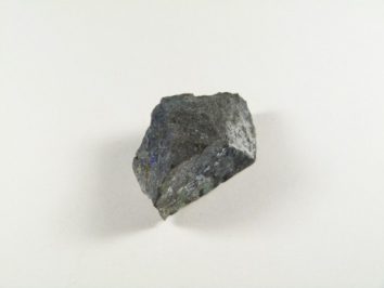 Calaghanit kamień. Szara, nieregularna bryłka minerału leżąca na jasnym tle.