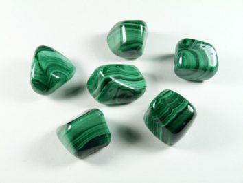 Malachit - kamień gładzony, polerowany, szlifowany i bębnowany w formie otoczaka. Sześć zielonych, wypolerowanych kamieni malachitu leży na białym tle.