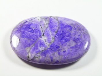 Howlit fioletowy - kamień gładzony, polerowany, szlifowany. Gładki, fioletowy kamień ozdobny o owalnym kształcie, leżący na białym tle.