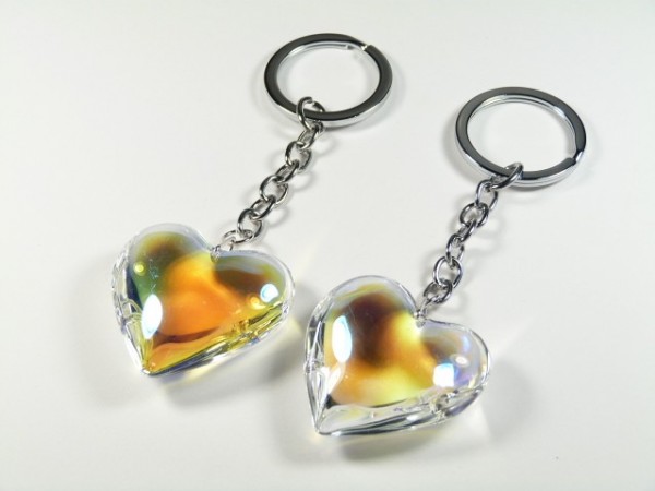 Dwa breloczki z przezroczystymi sercami na metalowych łańcuszkach, leżące na białym tle.