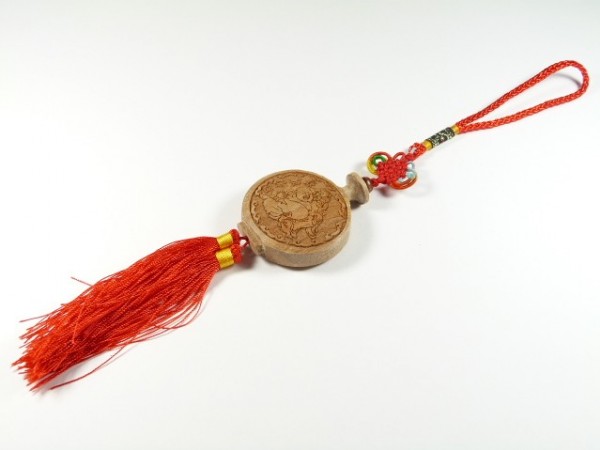 Chiński brelok z drewnianym medalionem i czerwoną frędzlą, leżący na białym tle.