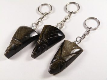 Trzy breloczki z rzeźbionymi głowami z ciemnego kamienia leżą na białym tle.