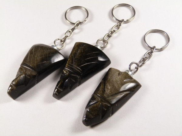 Trzy breloczki z rzeźbionymi głowami z ciemnego kamienia leżą na białym tle.