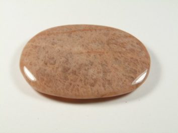 Kamień księżycowy - kamień gładzony, polerowany, szlifowany. Gładki, owalny kamień w odcieniu jasnego brązu leży na białym tle.