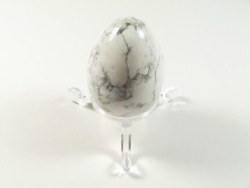Jajko howlit biały szlifowany. Jasny kamień w kształcie jajka z szarymi żyłkami, ustawiony na przezroczystej podstawce.