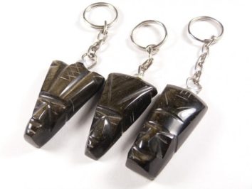 Trzy breloczki z ciemnego kamienia w kształcie egipskich masek na białym tle.