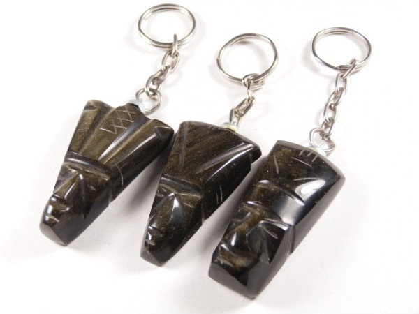 Trzy breloczki z ciemnego kamienia w kształcie egipskich masek na białym tle.