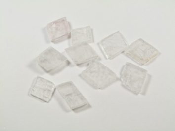 Spar islandzki - kamień naturalny, surowy, nieszlifowany. Kilka przezroczystych, kwadratowych kryształków leży na białym tle.