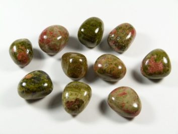 Unakit - kamień gładzony, polerowany, szlifowany i bębnowany w formie otoczaka. Dziesięć gładkich, polerowanych kamieni o zielono-czerwonych barwach na białym tle.