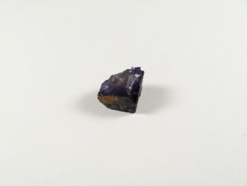 Fluoryt - kamień naturalny, surowy, nieszlifowany. Fioletowy, surowy kamień minerału leży na białym tle.