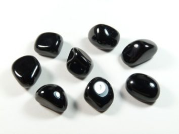 Onyks czarny - kamień gładzony, polerowany, szlifowany i bębnowany w formie otoczaka. Osiem polerowanych, czarnych kamieni leży na białym tle.