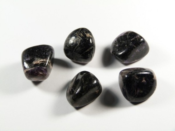 Czaroit - kamień gładzony, polerowany, szlifowany i bębnowany w formie otoczaka. Pięć polerowanych, ciemnych kamieni leży na białym tle.