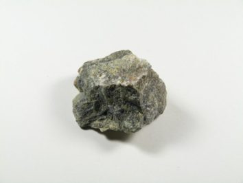 Serpentynit - kamień naturalny, surowy, nieszlifowany. Szara, nieregularna bryła kamienia leży na białym tle.