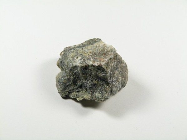 Serpentynit - kamień naturalny, surowy, nieszlifowany. Szara, nieregularna bryła kamienia leży na białym tle.