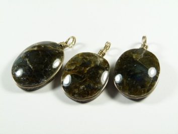 Trzy owalne wisiorki z ciemnych kamieni z metalowymi zawieszkami, ułożone na białym tle.
