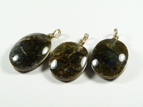 Trzy owalne wisiorki z ciemnych kamieni z metalowymi zawieszkami, ułożone na białym tle.
