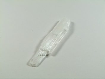 Biały, wydłużony kryształ leży na jasnym tle.