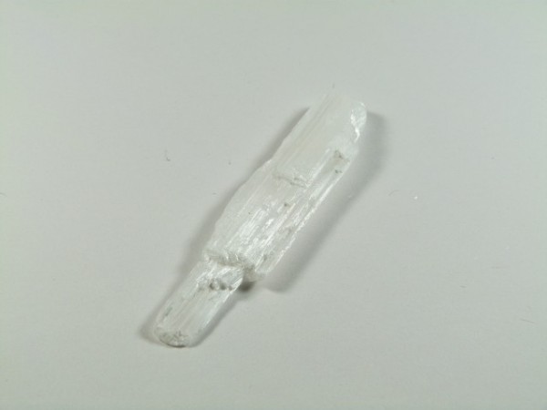 Biały, wydłużony kryształ leży na jasnym tle.