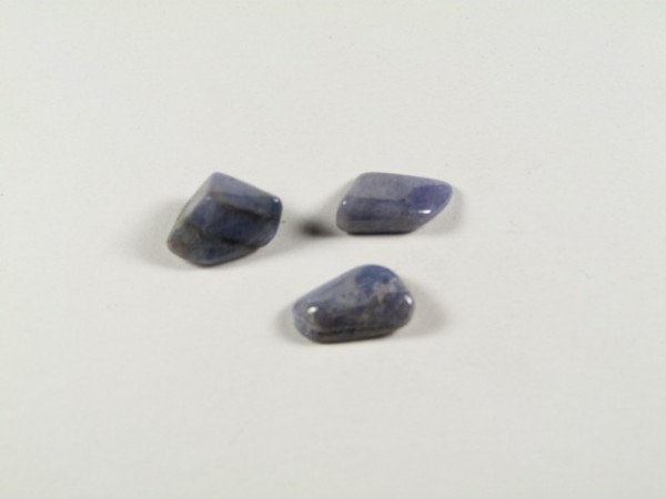 Tanzanit - kamień gładzony, polerowany, szlifowany i bębnowany. Trzy niebieskie, oszlifowane kamienie leżące na jasnym tle.