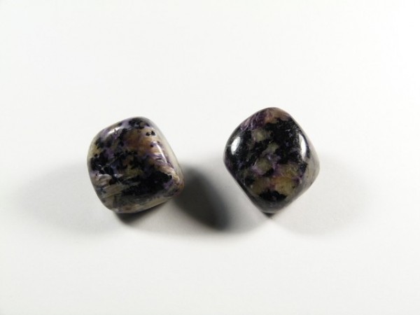 Czaroit - kamień gładzony, polerowany, szlifowany i bębnowany w formie otoczaka. Dwa polerowane kamienie o fioletowo-czarnych wzorach na białym tle.