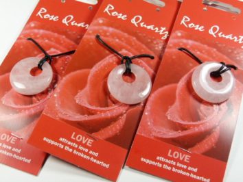 Trzy wisorki z różowego kwarcu na czarnych sznurkach, zapakowane na tle czerwonej róży.