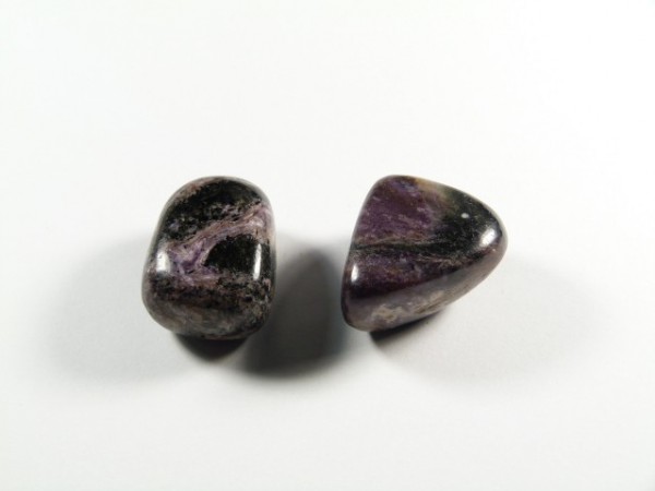 Czaroit - kamień gładzony, polerowany, szlifowany i bębnowany w formie otoczaka. Dwa wypolerowane, fioletowo-czarne kamienie leżą obok siebie na białym tle.