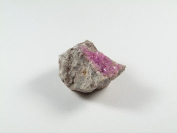 Kobaltokalcyt - kamień naturalny, surowy, nieszlifowany. Szara skała z widocznymi różowymi kryształkami na jednej stronie, na białym tle.