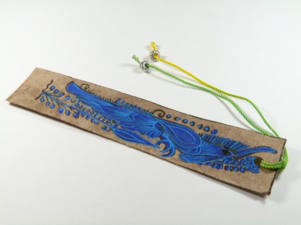 Ręcznie wykonana zakładka do książki z niebieskim rysunkiem ptaka i kolorowymi sznurkami.